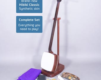 Puede incluir: Un juego completo de shamisen Hibiki Classic. El instrumento es de color marrón oscuro con una piel sintética blanca. El juego incluye una tela morada, accesorios y el texto "Brand-new Hibiki Classic Synthetic skin" y "Complete Set Everything you need to play!"