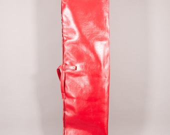Puede incluir: Una bolsa acolchada rectangular de color rojo brillante con un asa y una correa. La bolsa está hecha de un material brillante similar al cuero y tiene un cierre de cremallera. La bolsa está diseñada para guardar un objeto largo y estrecho.