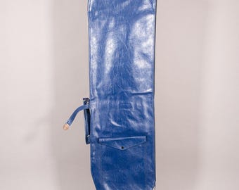 Puede incluir: Una bolsa de golf larga de vinilo azul con una superficie texturizada. La bolsa tiene un pequeño bolsillo con cierre de botón y un ribete rojo en la parte inferior. Una correa con hebilla está sujeta al lateral.