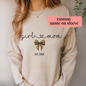 May include: Beige sweatshirt with the text "girl mom" in a script font, a leopard print bow, and the year "EST. 2026". The sleeve has the name "Mia" and a pink banner that says "custom name on sleeve".