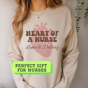 May include: Beige sweatshirt with the text "HEART OF A NURSE" above a pink anatomical heart illustration. Below the heart, the text reads "Labor & Delivery." A green rectangle with the text "PERFECT GIFT FOR NURSES" is at the bottom.