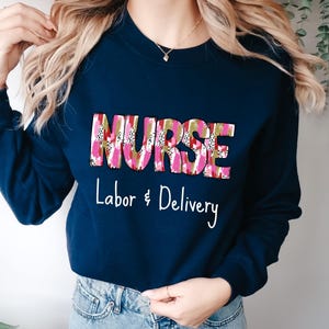 May include: Navy blue sweatshirt with the word "NURSE" in a pink, white, and gold patterned design. Below the word "NURSE" is the text "Labor & Delivery" in white script.