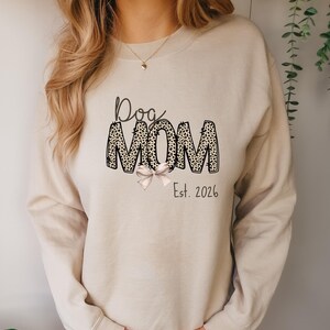 May include: Beige sweatshirt with the text "Dog MOM" in black script and leopard print. A light pink bow is below the word "MOM". The text "Est. 2026" is below the bow. The sweatshirt has long sleeves.