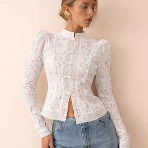 Puede incluir: Blusa de encaje blanco con cuello alto y mangas largas. La blusa presenta una parte delantera abotonada y un delicado estampado floral. La modelo lleva vaqueros azules. La blusa es una adición elegante a cualquier armario.