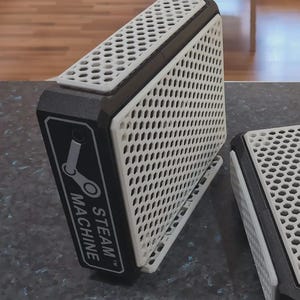 Könnte beinhalten: Zwei schwarz-weiße Steam Machine-Gehäuse. Die Gehäuse haben eine perforierte weiße Frontplatte und einen schwarzen Rahmen. Das Steam Machine-Logo ist auf der Vorderseite sichtbar. Ein Gehäuse steht aufrecht, das andere liegt flach.