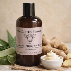 May include: A brown bottle of McCarthy Masons Tallow Milk Face Wash with a cow illustration on the label. The bottle is next to a small white bowl of cream, ginger root, and aloe vera leaves. The label says "Ginger & White Tea".
