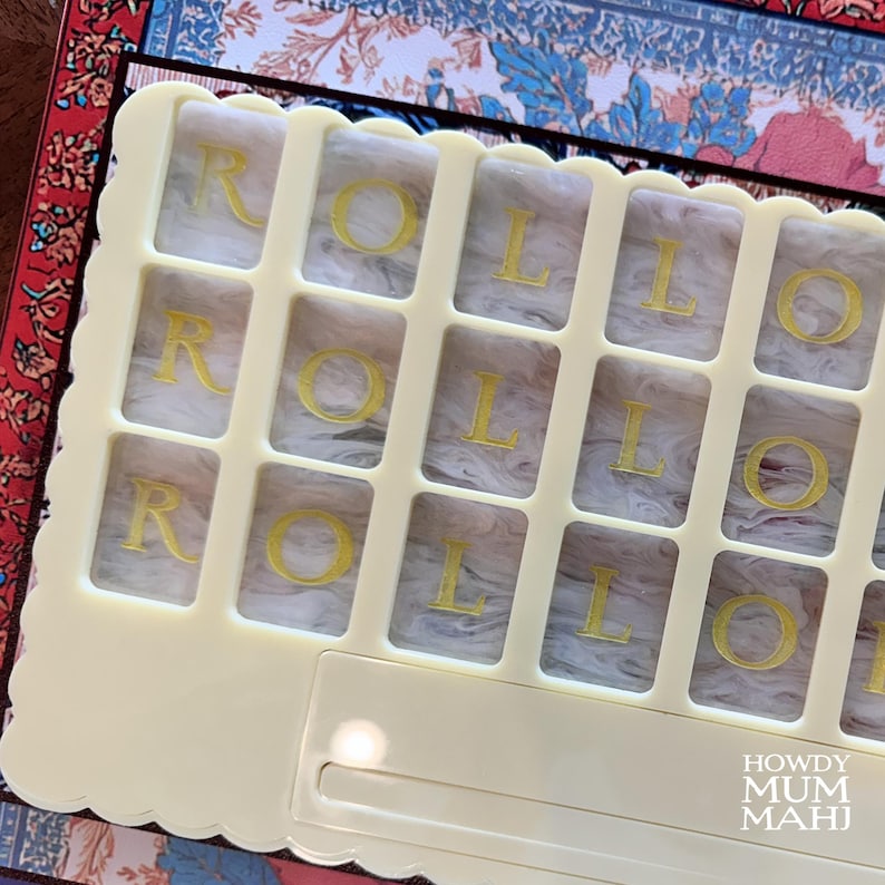 May include: A cream-colored game board with the word "ROLL" in gold letters. The board has a grid of rectangular spaces, each containing a letter. The background features a patterned design with floral and geometric elements. The text "HOWDY MUM MAHI" is visible.