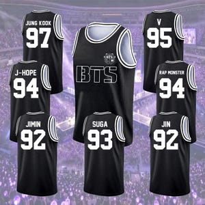 Puede incluir: Camisetas de baloncesto negras con detalles blancos y nombres y números de jugadores. Las camisetas muestran el texto "BTS" y los nombres "JUNG KOOK", "J-HOPE", "V", "RAP MONSTER", "JIMIN", "SUGA" y "JIN".