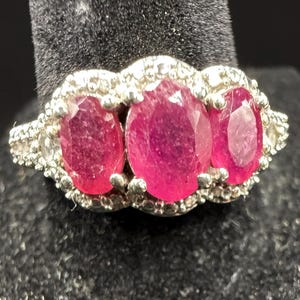 May include: A silver-toned ring featuring three oval-cut, deep pinkish-red gemstones arranged horizontally. The central stone is the largest, flanked by two smaller stones, all surrounded by a border of small, clear, sparkling accent stones.