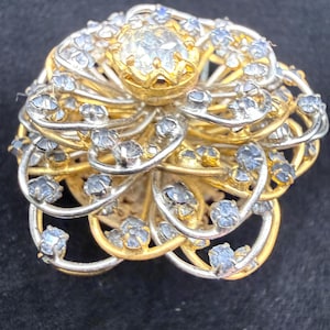 May include: A circular brooch featuring a layered, floral-inspired design with alternating gold and silver-toned metal wire loops. The center is set with a single clear stone, while the surrounding wire petals are accented with small, light blue stones.