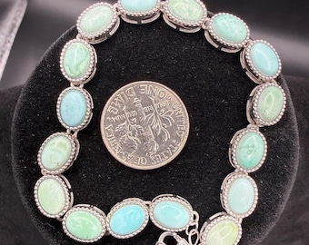 May include: A silver-toned link bracelet featuring fourteen oval-shaped, light blue and green cabochon stones set in beaded metal frames. The bracelet is displayed next to a United States dime for scale, showing the size of the individual stones.