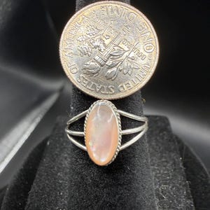May include: A silver ring featuring an oval-shaped, iridescent pinkish-white stone set in a twisted metal border with a triple-split band. A United States dime is positioned directly above the ring for scale, showing the relative size of the jewelry.