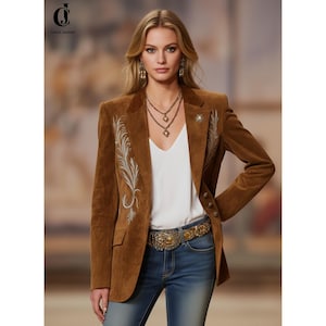 May include: A brown suede blazer with embroidered feather detailing. The blazer features a notched collar, three buttons, and a decorative pin. The model is wearing a white top, blue jeans, and a gold belt. The brand name "Corvis Jackets" is visible.