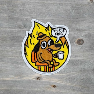 May include: Embroidered patch featuring a cartoon dog sitting in a chair, surrounded by flames. The dog is holding a mug and the speech bubble says "THIS IS FINE". The patch has a white border and is on a wooden background.