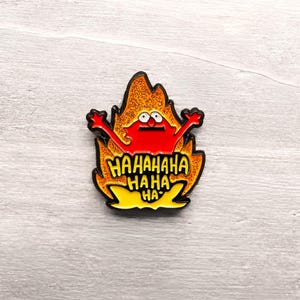May include: A metal enamel pin featuring a red character with wide eyes and raised arms centered within a stylized orange and yellow flame shape. The text HAHAHAHAHAHA HA is printed in black letters across the lower portion of the flames.