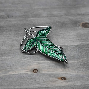 May include: A silver-toned leaf pendant with a vibrant green enamel finish. The leaf design features detailed veining and is set against a wooden background. A decorative piece of jewelry.