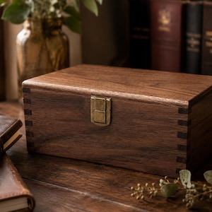 Peut inclure: Une boîte de rangement rectangulaire en bois de noyer brun foncé avec des motifs de grain visibles. La boîte présente des coins à joints carrés et un loquet en métal de couleur laiton antique centré sur le panneau avant.