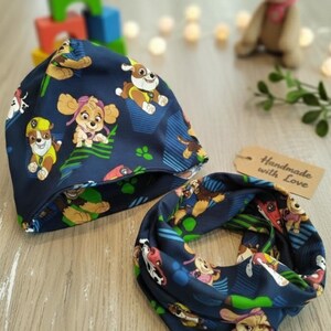 Könnte beinhalten: Ein marineblaues Mützen- und Halstuch-Set mit einem sich wiederholenden Muster von Cartoon-Welpen. Die Accessoires sind mit Bildern verschiedener Welpen in verschiedenen Posen verziert. Ein Etikett mit der Aufschrift "Handmade with Love" ist ebenfalls sichtbar.