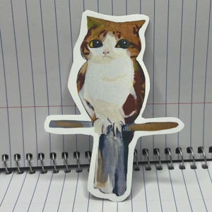 Peut inclure: Un autocollant représentant un chat perché sur un poteau en bois. Le chat a un pelage brun et blanc, avec une poitrine blanche et une tête brune. Le chat est assis droit, les pattes posées sur le poteau. Le fond est un carnet.