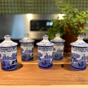 Könnte beinhalten: Fünf blau-weiße Keramik-Gewürzgläser mit Deckeln, beschriftet mit "Cinnamon", "Parsley", "Rosemary" und "Mixed Herbs". Jedes Glas zeigt eine malerische Illustration. Die Gläser sind auf einer Holzoberfläche angeordnet.