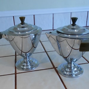 May include: Two matching chrome-plated sugar bowls with lids. Each bowl has a pedestal base, a curved body, and a dark brown handle. The lids are topped with a dark brown finial. The bowls are displayed on a tiled surface.