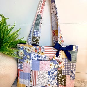 Puede incluir: Un bolso tote patchwork con una variedad de estampados florales, a rayas y de cuadros en tonos azules, rosas y blancos. El bolso tiene un lazo azul oscuro y asas a juego. El bolso está hecho de tela acolchada.