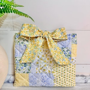 May include: A quilted square bag with a large bow. The bag is made of patchwork fabric in yellow, blue, and white floral and gingham patterns. The bow is made of the same floral fabric. The bag is likely a gift bag or decorative item.