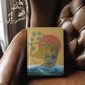 Pode incluir: Um diário de capa dura com uma capa dourada apresentando uma ilustração do rosto de um homem com um design geométrico e ondas do oceano. A obra de arte inclui detalhes em vermelho e azul, e o diário está em uma cadeira de couro marrom.