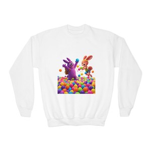 May include: White sweatshirt featuring a colorful graphic of two cartoon bunnies playing in a ball pit. One bunny is purple with a backpack, the other is orange. The ball pit is filled with various colored balls.