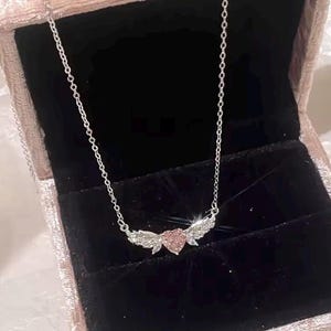 Może przedstawiać: Srebrny naszyjnik z r&oacute;żową zawieszką w kształcie serca i błyszczącymi skrzydłami. Naszyjnik jest prezentowany w r&oacute;żowym aksamitnym pudełku na biżuterię z czarnym wnętrzem. Łańcuszek jest delikatny, a zawieszka jest wyśrodkowana.