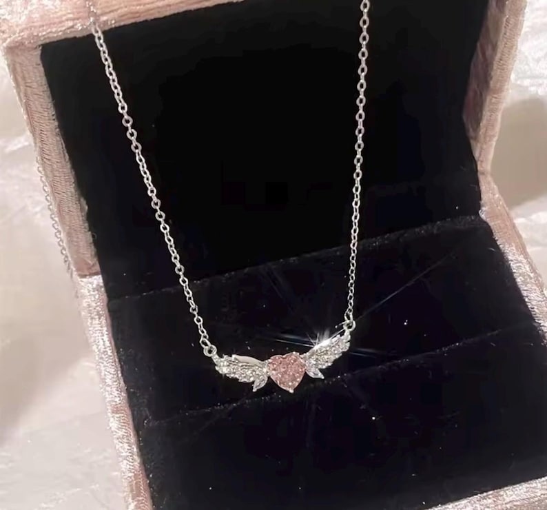 Może przedstawiać: Srebrny naszyjnik z r&oacute;żową zawieszką w kształcie serca i błyszczącymi skrzydłami. Naszyjnik jest prezentowany w r&oacute;żowym aksamitnym pudełku na biżuterię z czarnym wnętrzem. Łańcuszek jest delikatny, a zawieszka jest wyśrodkowana.