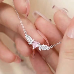 Może przedstawiać: Srebrny naszyjnik z r&oacute;żowym wisiorkiem w kształcie serca. Wisiorek otoczony jest skrzydłami, ozdobionymi małymi, błyszczącymi detalami. Naszyjnik jest zawieszony na łańcuszku.