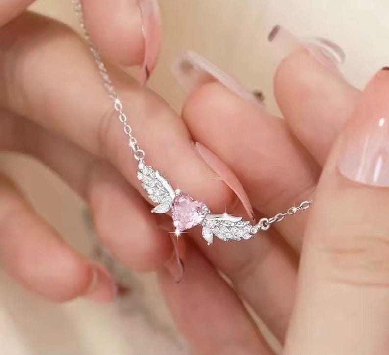 Może przedstawiać: Srebrny naszyjnik z r&oacute;żowym wisiorkiem w kształcie serca. Wisiorek otoczony jest skrzydłami, ozdobionymi małymi, błyszczącymi detalami. Naszyjnik jest zawieszony na łańcuszku.