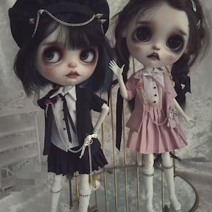May include: Two art dolls with large eyes and pale skin. One doll wears a black outfit with a chain and gun accessory, the other a pink dress. Both dolls have white knee-high socks and are displayed on a gold-toned stand.