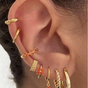 May include: An ear adorned with multiple gold earrings. The collection includes a twisted ear cuff, a sleek bar, a row of small orange-red gems, a textured stud, a floral drop earring, and several hoops, showcasing a variety of styles and designs.