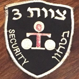 May include: A black and white security patch with Hebrew text and a central graphic. The patch has a shield shape with white text and border. The graphic is a stylized T-shape with circles.