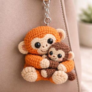 May include: A handmade crochet keychain featuring two adorable monkeys. The larger monkey is orange with beige accents, hugging a smaller brown monkey. The keychain is attached to a beige handbag with a gold-colored ring.