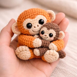 May include: Two crocheted monkey dolls, one larger and one smaller, are held in a hand. The larger monkey is orange with tan accents, and the smaller monkey is brown. Both have large black eyes and are smiling. The dolls are handmade.
