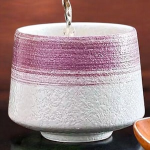 May include: A white teapot pours liquid into a small, handcrafted ceramic cup. The cup is white with a textured finish and a horizontal band of purple near the top. A small wooden spoon rests beside the cup, suggesting a tea ceremony.