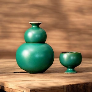 May include: A green ceramic sake set, comprising a double-gourd bottle and a small cup. The bottle and cup have a glossy finish and a gold rim. The set is displayed on a wooden surface.