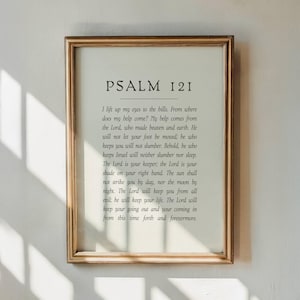 May include: A framed print with the text of Psalm 121 in black font. The frame is gold-colored and rectangular. The text is centered on a cream-colored background. The print is displayed on a white wall.