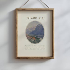 May include: Framed print featuring a landscape painting within an oval, with mountains and a river. The text "MICAH 6:8" is at the top, and a Bible verse is below. The frame is a light brown color, and the print is hanging by a string.