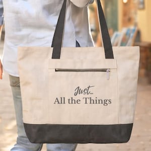 May include: A beige tote bag with black accents and the text "Just... All the Things" printed on the front. The bag has a zippered pocket and black handles. The bottom of the bag is black.