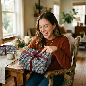 A delighted woman in a rust sweater laughs and points at the illustrated bookshelf pattern on her gift box, wrapped in navy book lover gift wrap with a burgundy ribbon bow, seated at a warm wooden dining table with candles and a ceramic mug nearby, the perfect bookish gift for the reader in your life.