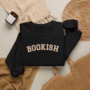 Pode incluir: Uma camisola preta de manga comprida com a palavra BOOKISH num tipo de letra universitário arqueado de cor dourada no peito. A peça tem um decote redondo, punhos canelados e uma bainha canelada, com um design atlético clássico e simples.