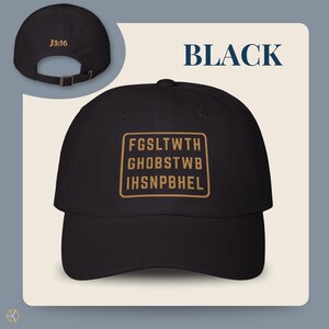 Black baseball cap with the text "BLACK" and "J3:16". The front of the cap features a gold rectangular design with the text "FGSLTWTH GHOBSTWBI HSNPBHEL". The cap has an adjustable strap.