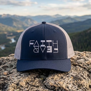 May include: A navy blue trucker hat with a grey mesh back and a solid front panel. The front features white embroidered text that reads FAITH over FEAR, with the word FEAR printed upside down to create a mirrored, symmetrical design.