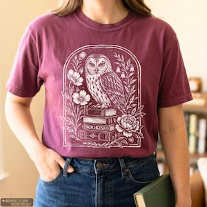 Pode incluir: Uma t-shirt de manga curta cor de vinho com uma impressão gráfica branca de uma coruja pousada numa pilha de livros. O design é emoldurado por um arco de flores e folhagem, com a palavra BOOKISH impressa na lombada de um livro.