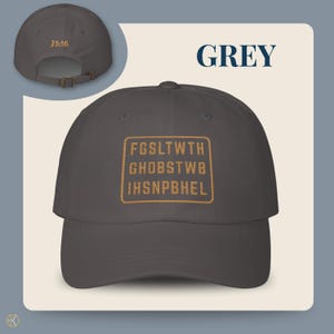 Grey baseball cap with the text "GREY" and "J3:16". The front of the cap features a gold rectangular design with the text "FGSLTWTH GHOBSTWBI HSNPBHEL". The cap has an adjustable strap.