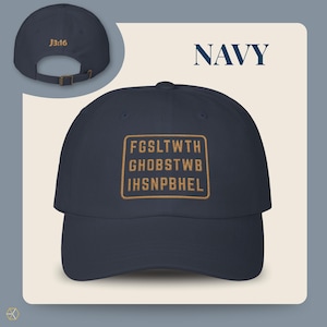Navy baseball cap with the text "NAVY" and "J3:16". The front of the cap features a gold rectangular design with the text "FGSLTWTH GHOBSTWBI HSNPBHEL". The cap has an adjustable strap.
